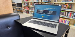 An open laptop logged into Windows 11 sits on a black table next to a cushioned chair. Shelves of teen books are in the background.