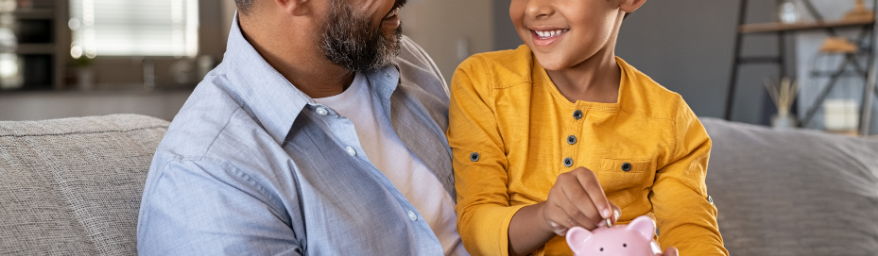 Money as You Grow page header Adult man and young boy smile while putting a coin in a pink piggy bank.