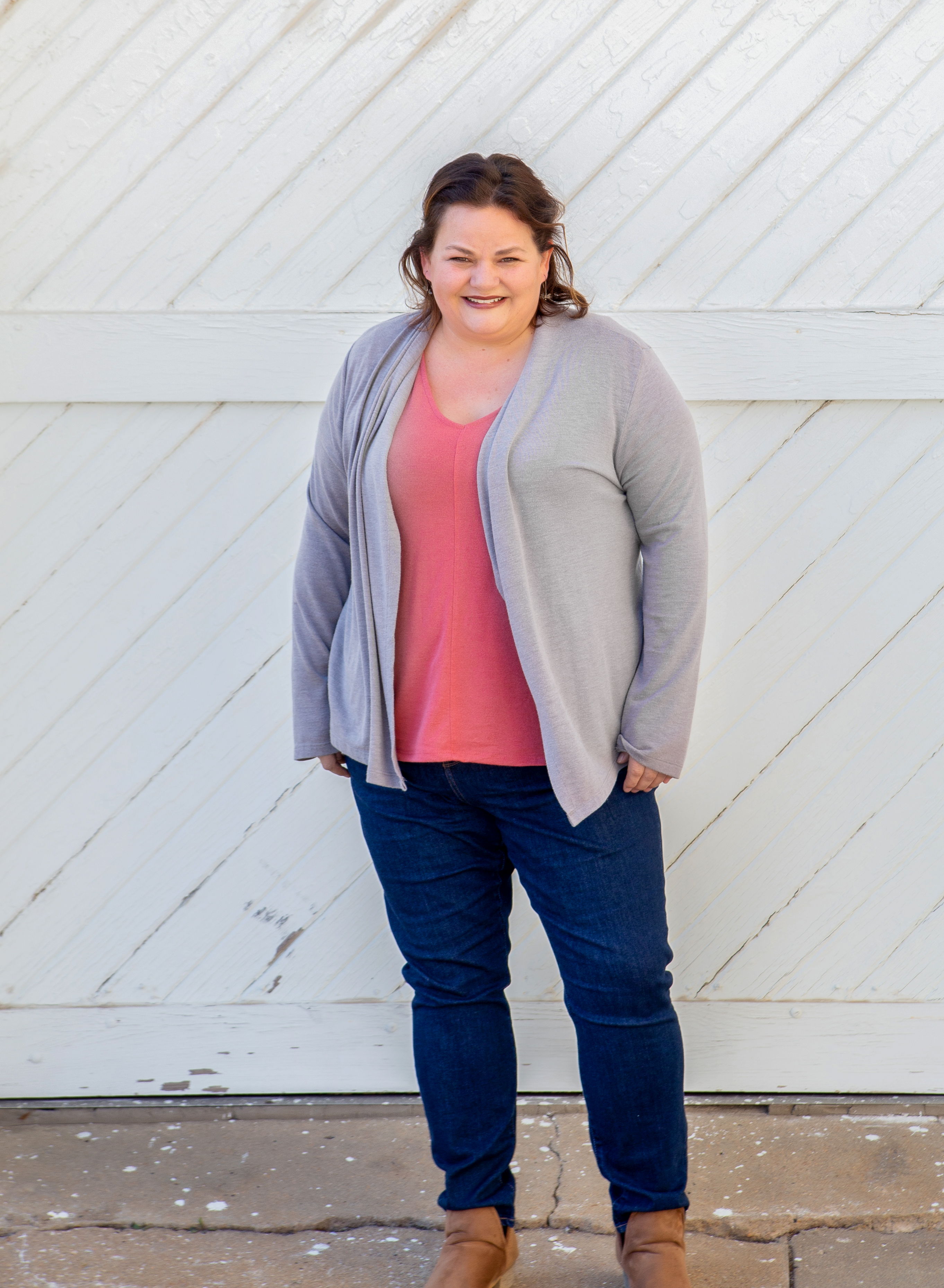 Standing in front of a white wood wall is Rianne, heavy set white woman with wavy hair pulled back wearing jeans, a pink top and a grey cardigan with brown booties