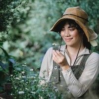 woman with dark, curly hair wearing a straw hat holding a small white flower in a garden