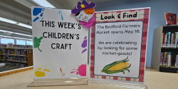 Fun Activities Two signs stand on a counter, one displaying a completed weekly foam children’s craft and the other inviting kids to participate in a Look and Find activity on the Children’s floor for a prize.