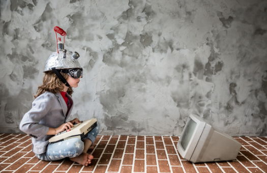 tween girl sitting on a brick floor with a colander covered in foil and an antenna wearing goggles, her legs are crossed and there is an 80's style monitor in front of her with a keyboard in her lap