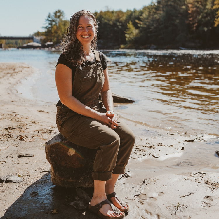 Woman, dressed in brown overalls and sandals, sitting on rock by a river