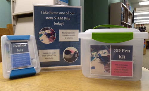 Two plastic containers and a sign displayed on a table in the library's children and teen area. One is a small, square plastic tub with a sign saying Ozobot kit. A sign in the middle says Take home one of our new STEM kits today, and another plastic tub with a sign. Second container is a 3D Pen kit.