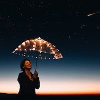 woman with short, dark hair under an umbrella made of lights, looking up at the night sky