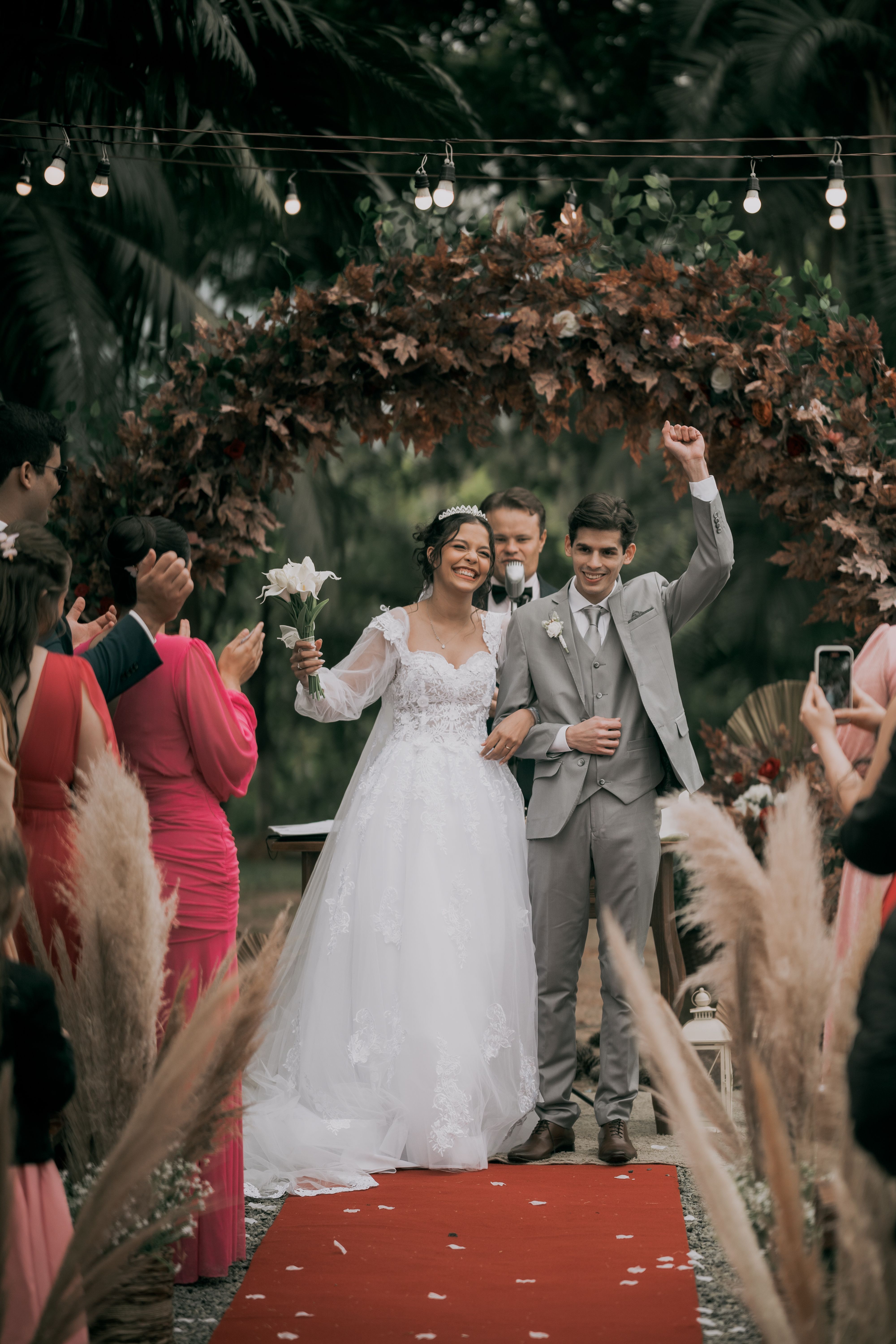 A smiling bride and groom exit their outdoor wedding ceremony while guests applaud.