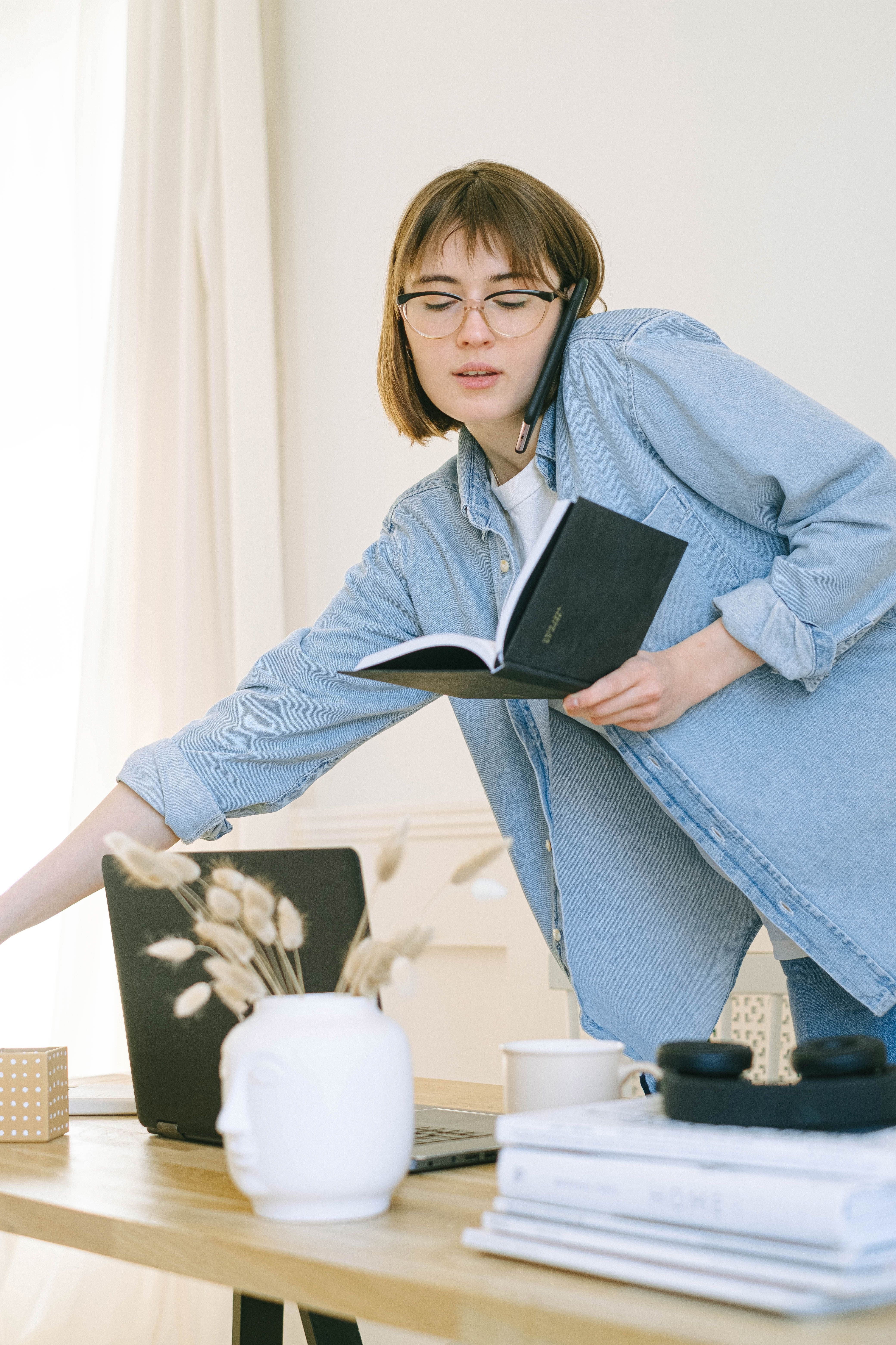 Courtesy of ivan-samkov from pexels Young woman in a denim button down talking on her phone while consulter her paper calendar while leaning over her desk.