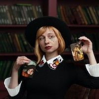 woman with auburn hair in a black hat and top with white cuffs holding tarot cards