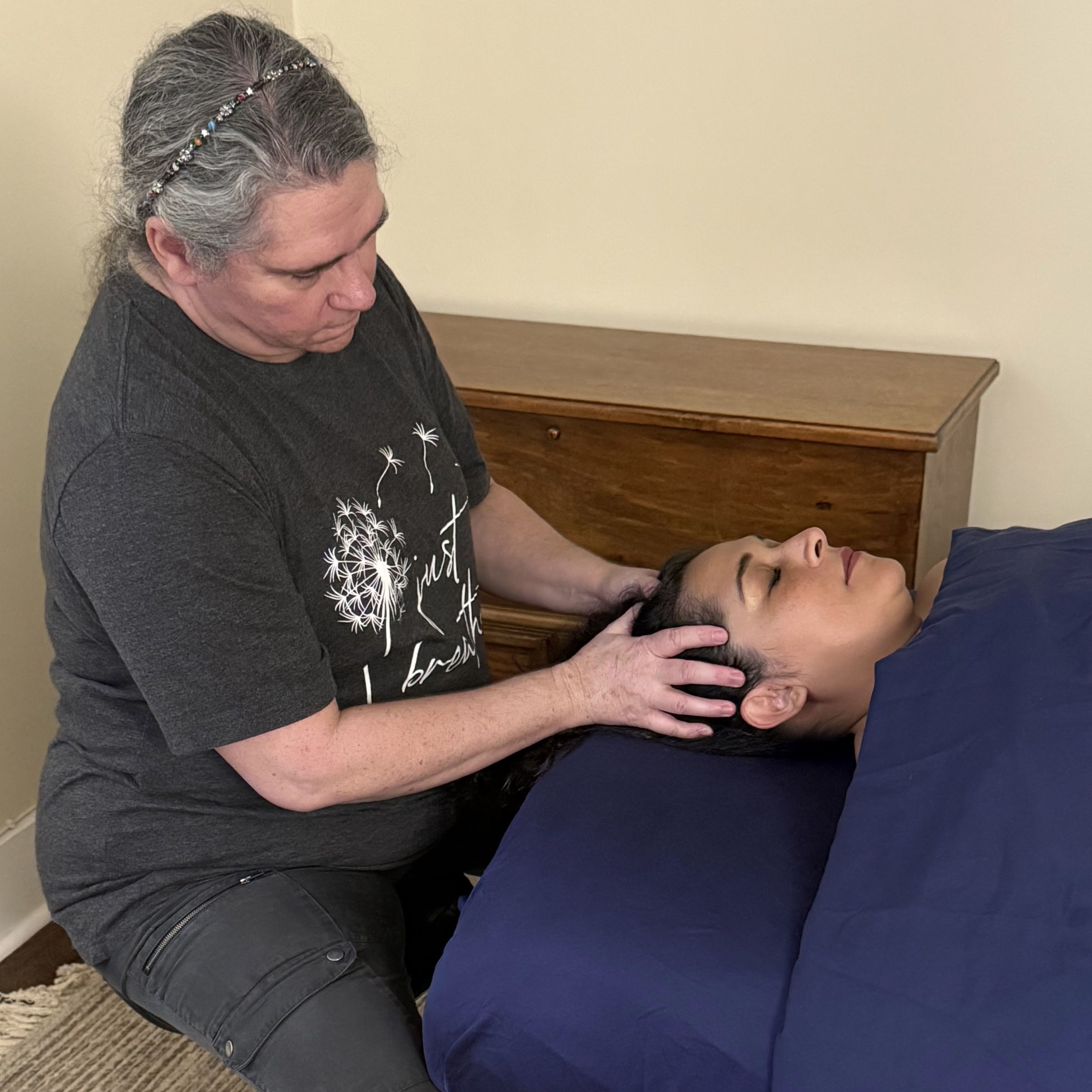 What Matters Renee sitting at the head of the massage table, looking at her hands on her client's scalp. The client has dark hair and brown skin and her eyes are closed.