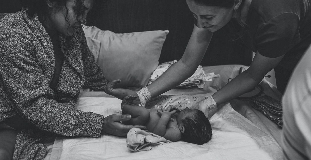 Black and white photo of newborn lying on bed, while checked on by midwife and mom