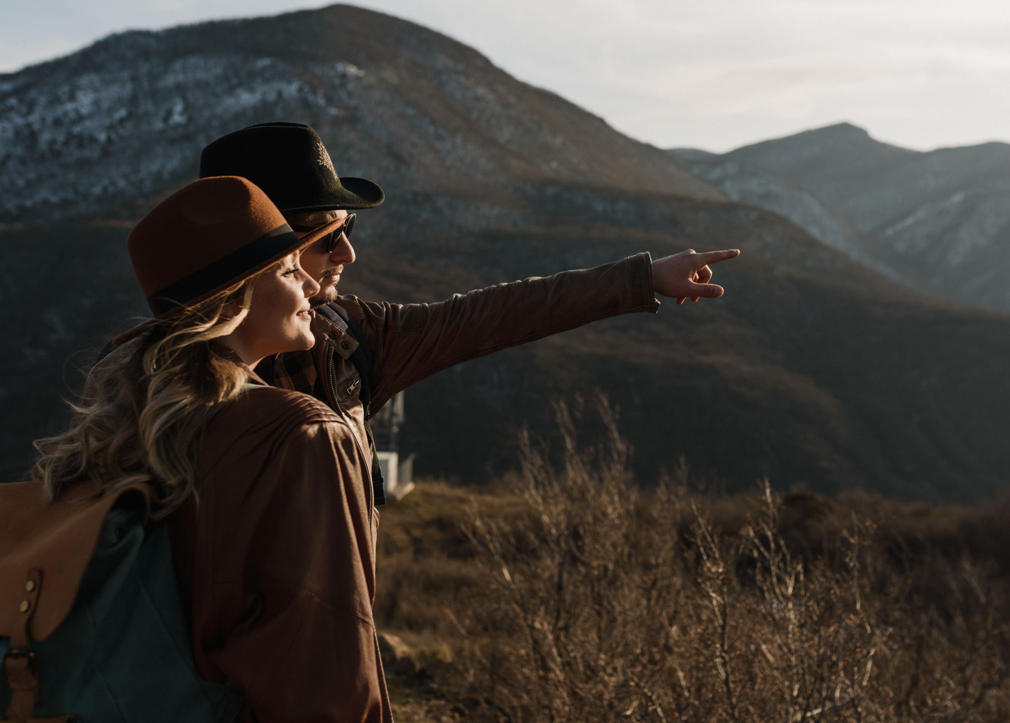 Two people dressed in long sleeves and hats, looking off to the right, with mountains in the distance. The person farther aways pointing to something in the distance.