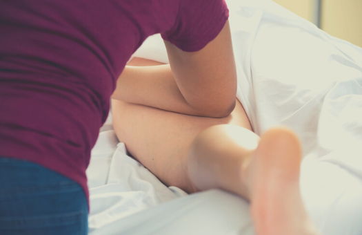 A white massage therapist in blue pants and a maroon top with their forearm and elbow in the back of an exposed white leg. The rest of the body is covered with a white sheet and they are laying on a white sheet with a pillow under their foot