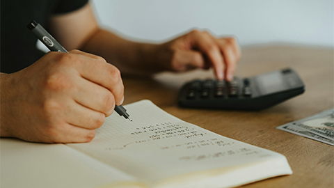 desk surface with a noteboook and calculator and a person writing