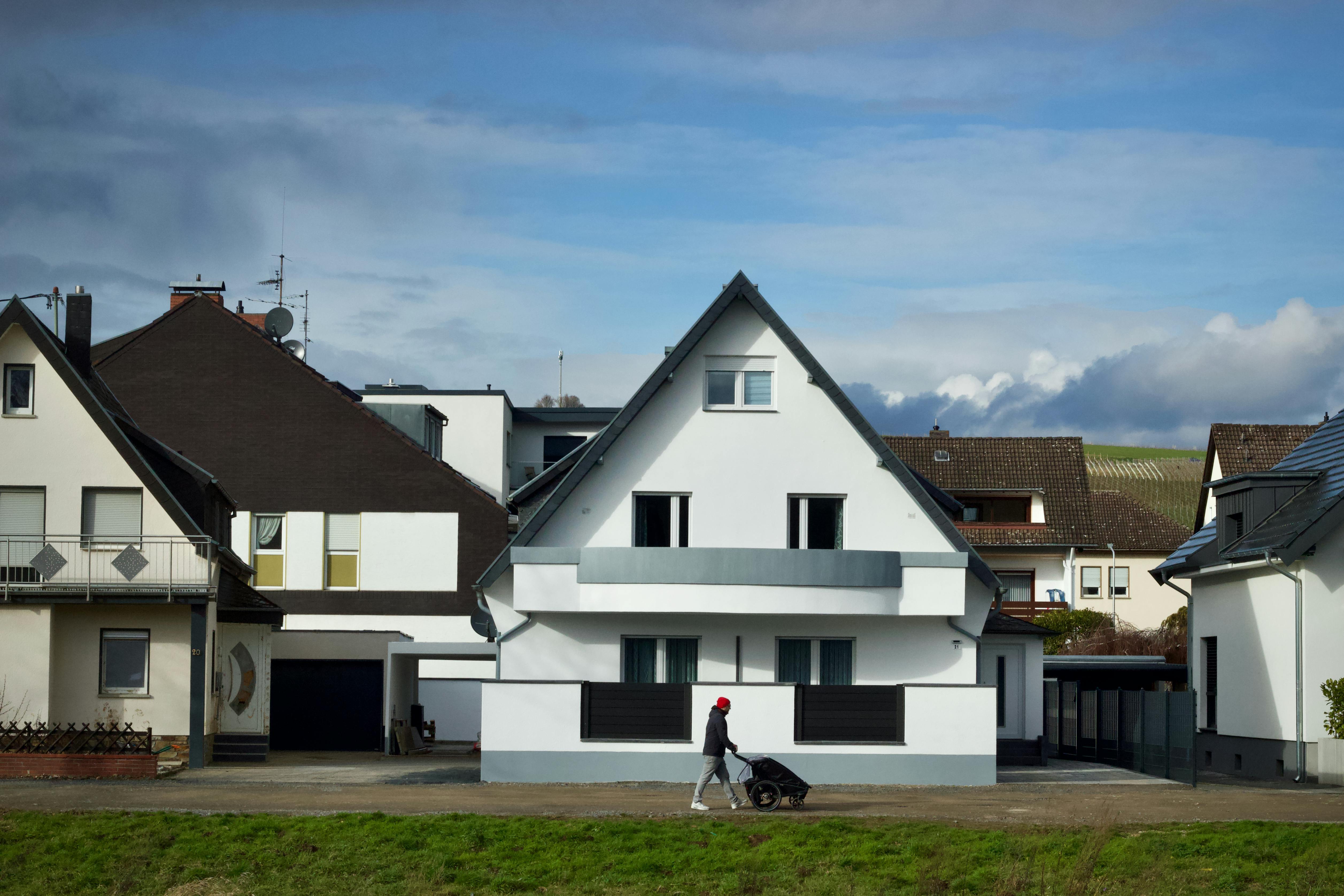 A person with a stroller walks by contemporary suburban houses under a bright sky.