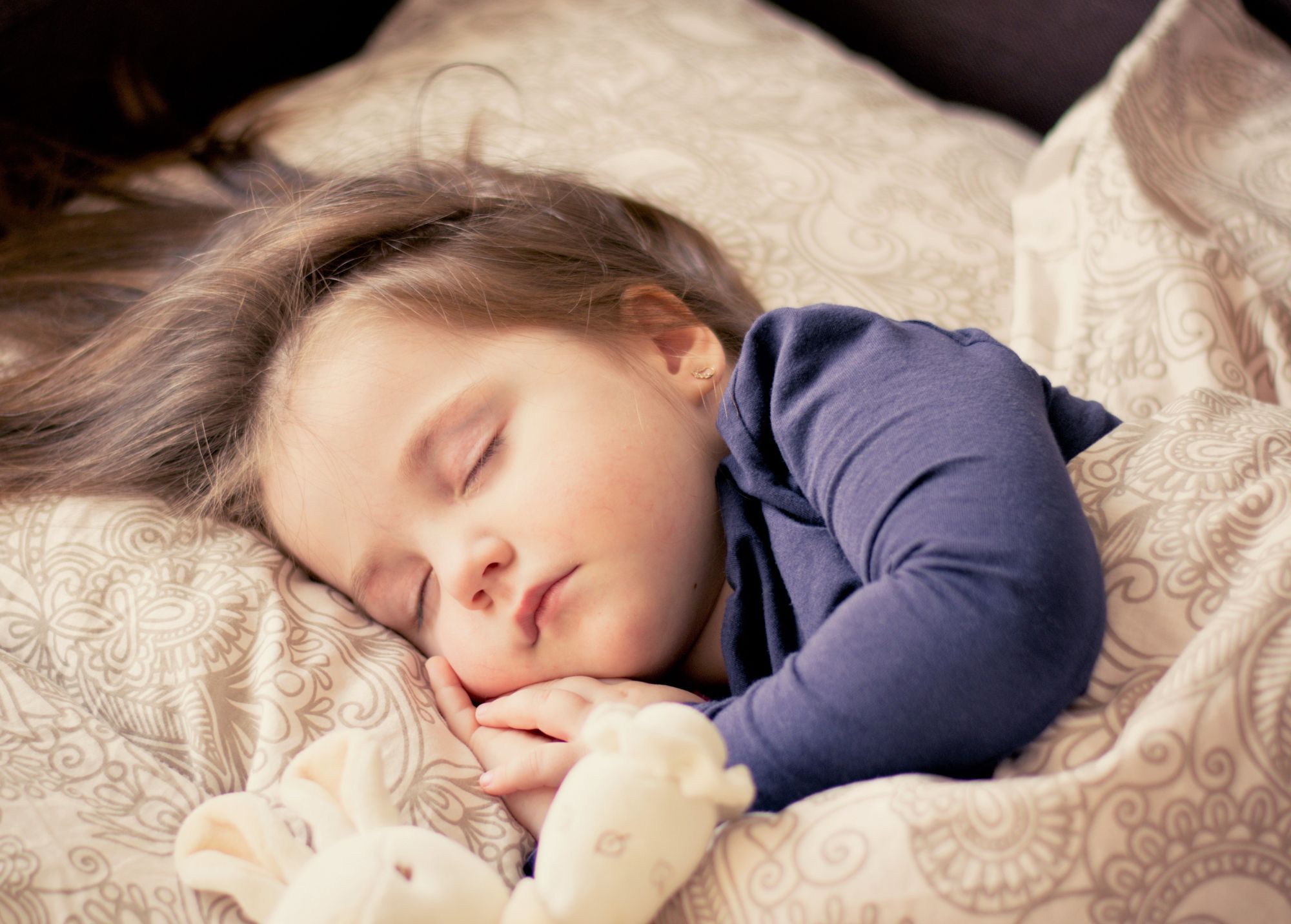 A small child sleeping on pillow, wearing a long-sleeved blue shirt with hands  a stuffed toy with hands folded under her cheek and a stuffed toy nearby