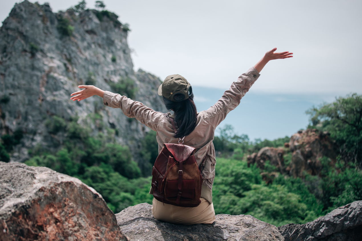 woman sitting on a rock facing a ravine full of trees with her arms in the air