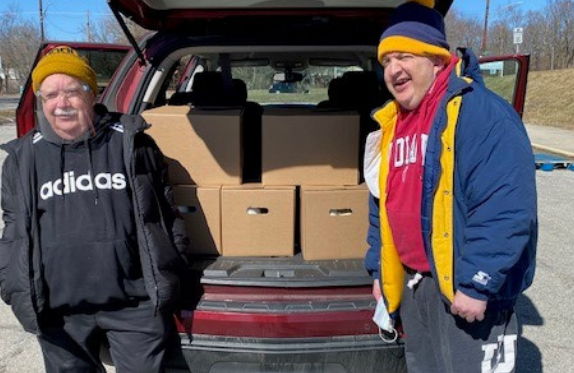 two older men stand on either side of a vehicle with it's liftgate open, full of boxes.