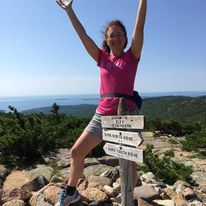 Woman on top of mountain, at trail sign with hands raised about her head