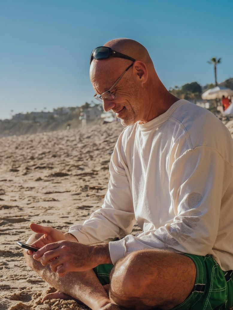 man on beach smiling while using smartphone