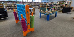 Play Area An open floor space includes a giant Connect Four game, a table-top train set, two tables with chairs, two cushioned armchairs, shelves of small toys and kits, and a giant foam log. Shelves of children’s books surround the space.
