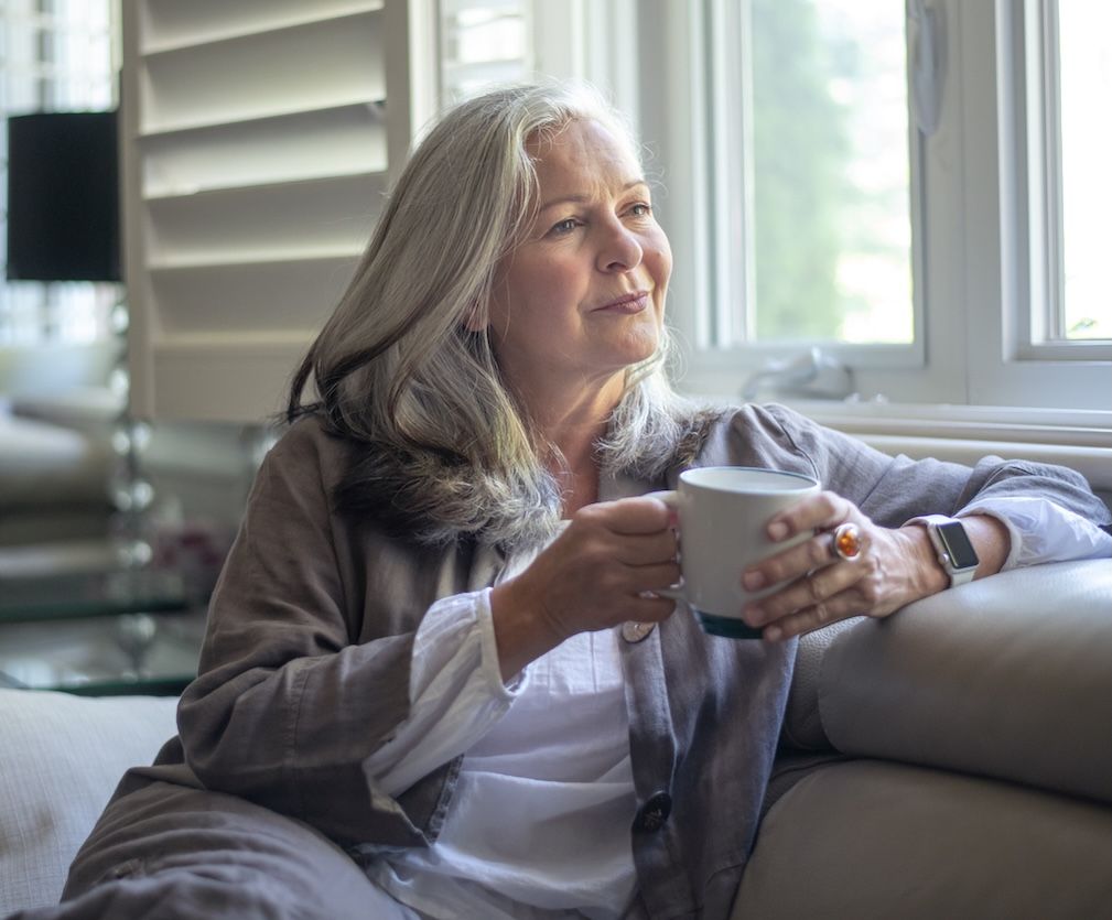 mature woman sitting on couch drinking coffee and looking out windows