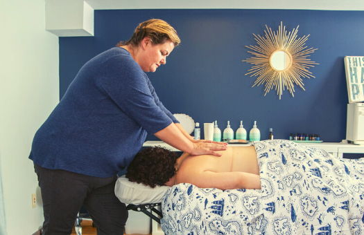 Rianne, a fat, white woman wearing a blue top and grey pants, leaning over a white woman giving a massage on her exposed back
