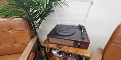A brown retro-style record player sits on a faux wood and metal side table between two brown vinyl chairs. Stacks of record album covers are displayed in the table underneath.