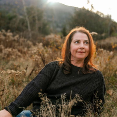 white woman with shoulder length brown hair sitting on ground with long grass